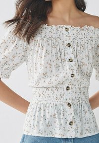 Hvid blomstret bluse med korte puffærmer, samlet smock-talje og knapper foran. Har små, brune blomster mønstre på stoffet.
