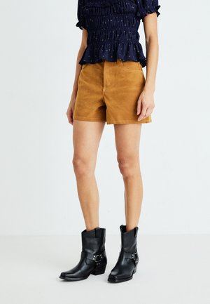 Vrouw in bruine suède shorts, zwarte leren laarzen tot halverwege de kuit, en een marineblauw gerimpeld topje tegen een effen witte achtergrond.