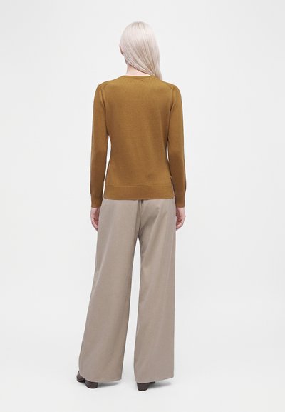 Femme aux cheveux blond platine porte un pull marron moutarde et un pantalon large beige, debout dos à l’objectif sur un fond blanc uni.