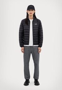 Veste matelassée noire avec une fermeture éclair à l'avant, t-shirt gris en dessous, jean gris et chaussures noires. Une casquette noire complète la tenue.