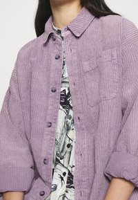 Lavendel cordjacka med knappfront, krage och ficka. Mjuk textur med vertikal ribb och uppvikta ärmar.