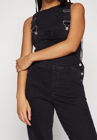 Jeune personne portant une salopette en jean noire et un top noir sans manches, debout, un bras croisé sur l'autre.