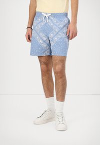 Mann iført lys blå mønstrede shorts med hvit snøring, hvite joggesko og hvite sokker, stående mot en ensfarget bakgrunn.
