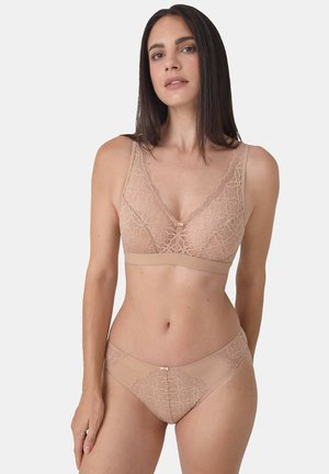 Frau trägt einen hautfarbenen Spitzen-BH und passende Panties, steht vor einem schlichten weißen Hintergrund und schaut nach vorne.