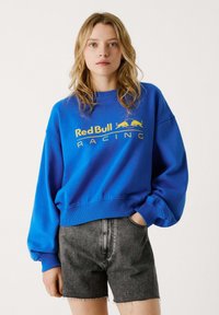 Plava dukserica s okruglim izrezom i rebrastim manžetama, sa žutim logotipom "Red Bull Racing". Nosena s tamno sivoj traper šortsevima.