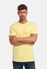 T-shirt en coton jaune avec un col rond, des manches courtes et un petit logo blanc sur la poitrine, associé à un jean en denim bleu.