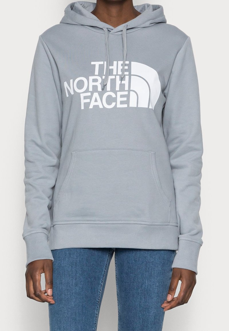 Personne portant un sweat à capuche gris clair The North Face avec une poche avant et un jean bleu, debout devant un fond uni.