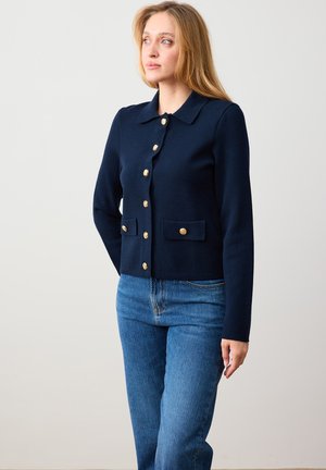 Kobieta ubrana w granatowy kardigan na guziki z złotymi guzikami oraz niebieskie dżinsy, stojąca na tle jednolitego, jasnego tła.