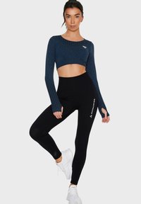 Marineblaues, kurz geschnittenes Langarmoberteil mit Rippdetail und schwarze, hochgeschnittene Leggings mit dem Schriftzug "SQUATPROOF", kombiniert mit weißen Sportschuhen.