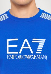 Modrá športová košeľa s bielym logom "EA7 EMPO". Má okrúhly výstrih a pruhované ramenné akcenty. Vyrobená z hladkého, elastického materiálu.