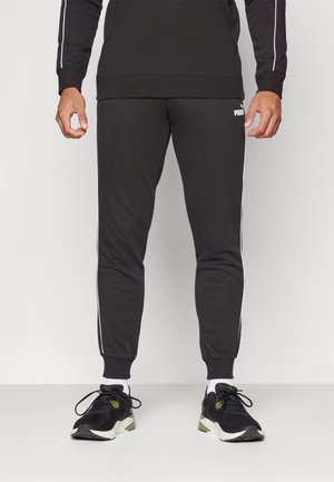 Schwarze Jogginghosen mit weißen Seitstreifen, taillierter Schnitt, weicher Stoff und elastischen Bündchen. PUMA-Logo am linken Oberschenkel. Getragen mit schwarzen Sneakers.