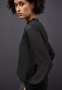 Top negro de manga larga con textura en chevrón, mangas transparentes y cuello con volantes. La tela parece ligera y suave.