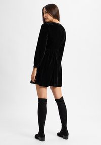 Schwarzes Samtkleid mit langen Ärmeln, tailliertem Schnitt und A-Linien-Rock. Kombiniert mit kniehohen schwarzen Stiefeln aus Stoff und einer strukturierten Oberfläche.