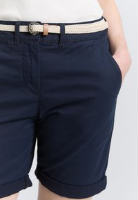 Marineblaue Baumwollshorts mit umgeschlagenen Säumen, ausgestattet mit einem gewebten beigen Gürtel und einer silbernen Metallschnalle. Sichtbare Seitentasche und glatte Textur.