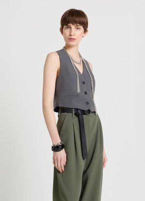 Gilet grigio senza maniche con scollo a V e tre bottoni, abbinato a pantaloni verdi a vita alta e cintura nera; dettaglio di collana a catena d'argento.