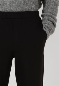Svarta byxor med elastisk midja och sidofickor, slät textur och en figursydd design. Bärs med en ljusgrå, fluffig stickad tröja.
