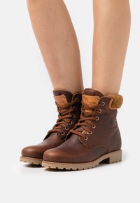 Bruna läderankelstövlar med en vadderad krage, snörning och en robust sula. Har en logotyp-patch och synliga sömsdetaljer.