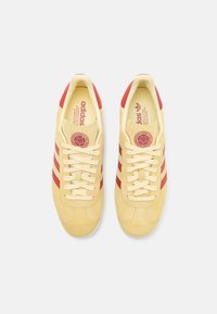 Zapatillas de ante amarillas con rayas y acentos rojos, que cuentan con un diseño de cordones y un interior acolchado. Logo impreso en la lengua.