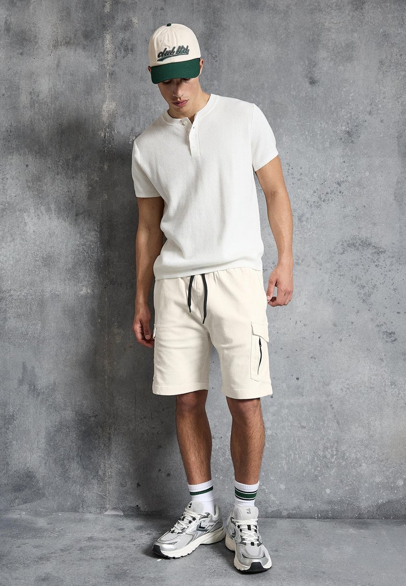 Junger Mann, der ein weißes Henley-Shirt mit kurzen Ärmeln, cremefarbene Cargoshorts, weiße Turnschuhe, gestreifte Socken und eine grün-weiße Baseballkappe trägt und vor einer grauen Wand steht.