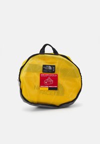 Borsa a rete gialla con accenti neri, dotata di maniglia superiore e di un'etichetta rossa che mostra le informazioni sul prodotto e un design grafico.