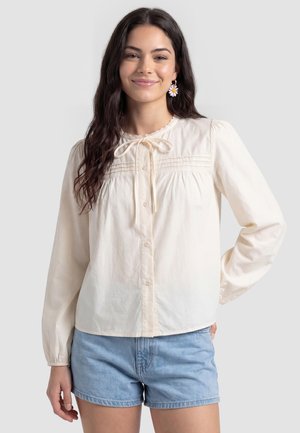 Jonge vrouw met lang donker haar draagt een crèmekleurige blouse met knopen en strik, en lichtblauwe denimshorts, glimlachend met hand op de heup.