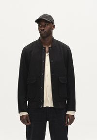 Črna volnena bomberska jakna z dvema sprednjima žepoma, gumbi na zapenjanje, rebrastimi manšetami in usklajeno črno kapo. Model nosi pod jakno kremasto belo srajco.