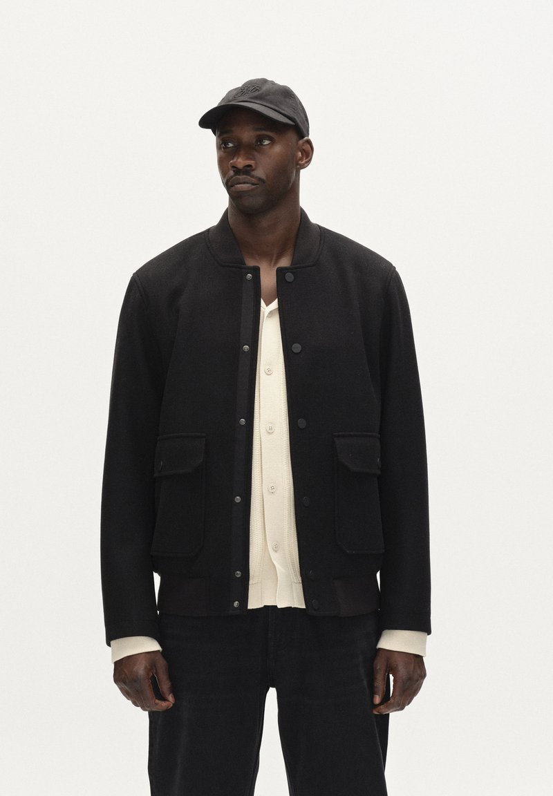 Črna volnena bomberska jakna z dvema sprednjima žepoma, gumbi na zapenjanje, rebrastimi manšetami in usklajeno črno kapo. Model nosi pod jakno kremasto belo srajco.