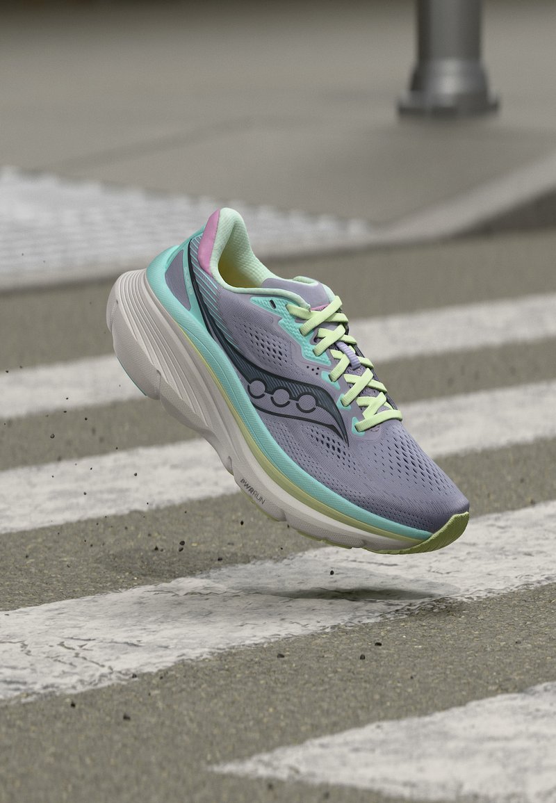 Chaussure de course violet clair avec des accents vert menthe et rose, flottant au-dessus d'un passage piéton avec du petit gravier volant en dessous.