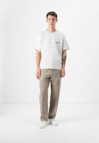 Camiseta gris de manga corta con un logo negro, combinada con pantalones beige claros y zapatillas blancas. La tela parece suave y informal.