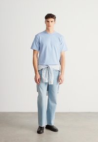 Világoskék, kerek nyakú póló világos farmernadrággal párosítva. Csíkos ing van megkötve a derékon. Helyi tervezésű fekete cipők egészítik ki a megjelenést.