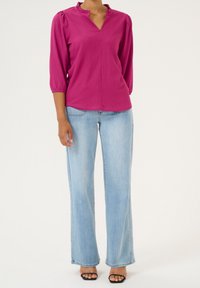 Blouse fuchsia avec un col à volants et des manches trois-quarts, associée à un jean large bleu clair et des sandales noires à bouts ouverts.