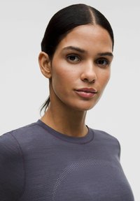 Tumeda juustega naine on üles pandud, seljas tumedat halli sobivat toppi, millel on peeneline täppide tekstuur, heleda ühtlase tausta ees.