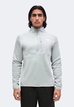 Homme aux cheveux noirs portant une veste polaire gris clair à zip quart, avec une poche poitrine zippée et un pantalon noir, debout devant un fond blanc.