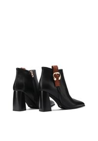 Stivali ankle in pelle nera con punta appuntita, tacco spesso, zip laterale e dettaglio di fascia marrone con fibbia dorata.