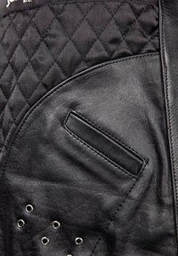 Schwarze Lederjacke mit einer strukturierten Rautensteppung, einer flachen Tasche und silbernen Beschlägen mit Ösensegmenten entlang des unteren Randes.