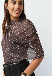 Brown polka dot sheer wrap, featuring white dots, draping around the shoulders; paired with black high-waisted pants and a black belt.