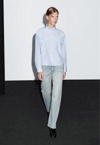 Camicia button-up a righe blu chiaro con due tasche sul petto, abbinata a jeans azzurri a gamba dritta e scarpe nere lucide.