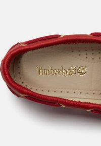 Άνω όψη μπεζ πάτου με χρυσό λογότυπο Timberland μέσα σε κόκκινο δερμάτινο μοκασίνι με ραμμένες λεπτομέρειες και στρογγυλές οπές.