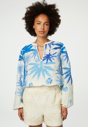 Langärmlige, leichte Bluse mit blauen Blumenmustern auf beigem Stoff; hat einen Schlüsselloch-Ausschnitt und eine lockere Passform, getragen mit hellen Shorts.