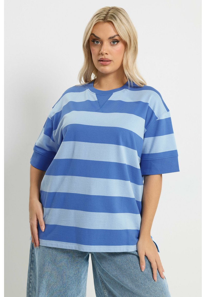 Femme blonde portant une chemise à manches courtes surdimensionnée à rayures bleues et bleu clair et un jean bleu clair, debout devant un fond blanc.