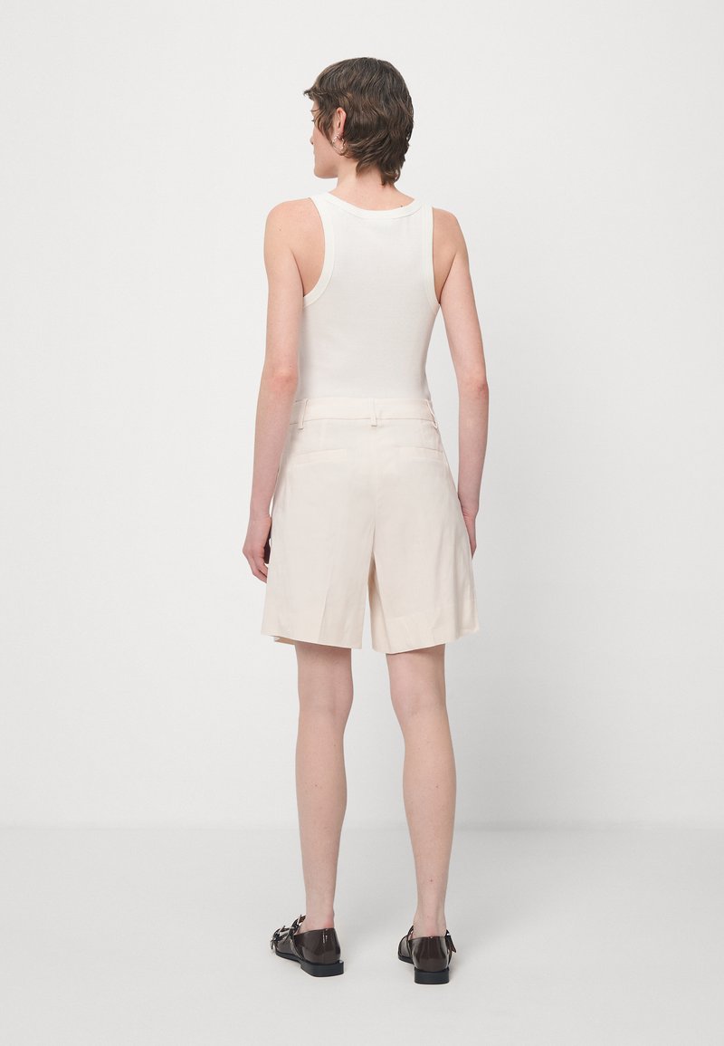 Beige Leinenshorts mit taillierter Passform, zwei Gesäßtaschen und umgeschlagenem Saum. Kombiniert mit einem weißen ärmellosen Oberteil und dunklen Schuhen.