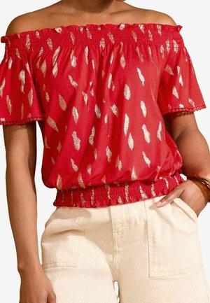 Femme portant un chemisier rouge épaules dénudées avec des taches dorées brillantes et une taille élastique, associé à un pantalon beige clair taille haute.