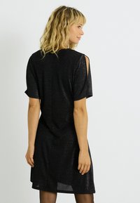 Robe noire avec une finition scintillante, manches courtes, découpes aux épaules et coupe décontractée. Présente un ourlet arrière légèrement plus long.