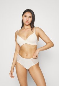 Ljusbeige bralette med V-ringning, tunna band och sömlös design; kombinerad med högmidjade trosor med en genomskinlig paneldetalj.