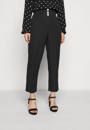 Kvinde iført sorte bukser med høj talje og tre guldknapper samt sorte hæle med ankelrem, kombineret med en sort- og hvidprikket bluse.