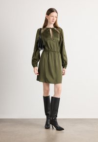 Olijfgroene jurk met lange pofmouwen, kantdetails bij de schouders, een strik in de taille en een keyhole-nekline. Gepaarde met zwarte kniehoge laarzen.