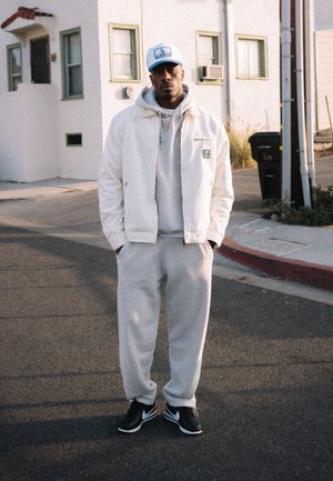 Veste blanche sur un sweat à capuche gris et un pantalon de survêtement, complétée par des baskets noires. La veste arbore un patch logo vert et a une coupe décontractée.