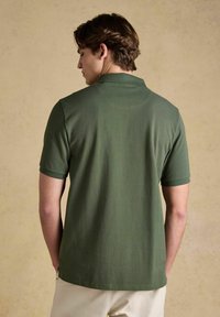 Mężczyzna stojący tyłem, ubrany w oliwkowozieloną koszulkę polo z krótkim rękawem oraz beżowe spodnie na jednolitym beżowym tle.