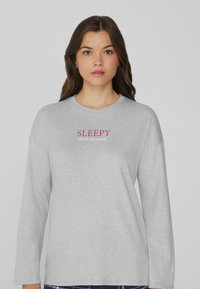 Camisa de manga larga gris hecha de un tejido suave que presenta la palabra "SLEEPY" en rosa y "state of mind" en un texto más pequeño debajo. Diseño simple y relajado.