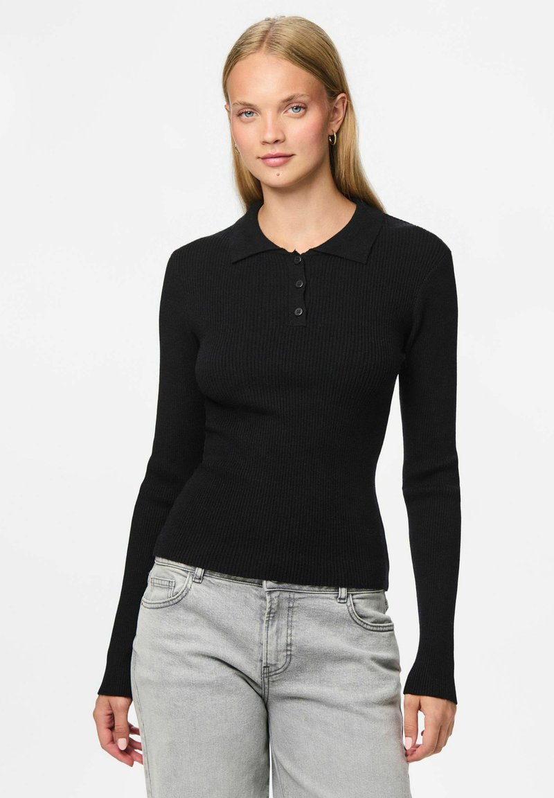 Plava žena u pripijenom crnom rebrastom dukserici dugih rukava i svijetlosivim trapericama, stoji naslonjena na bijelu pozadinu.